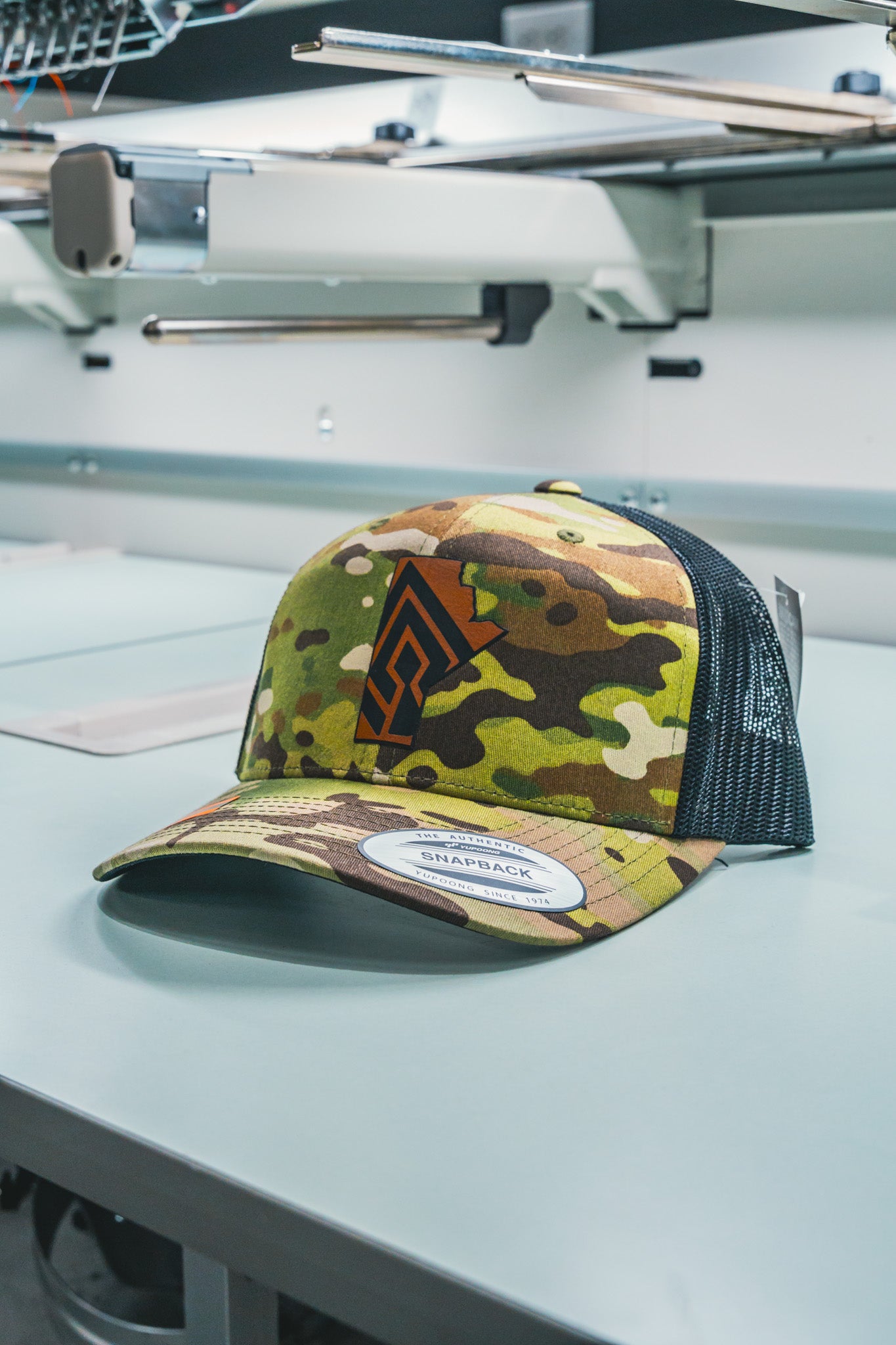 A camo snapback cap featuring a bold logo on the front, displayed in a shop setting.